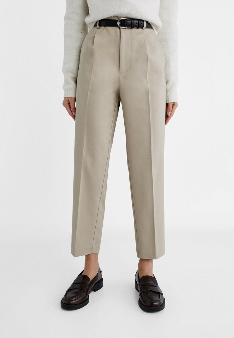 Pantalon beige taille haute, cintré, avec des plis sur le devant, porté avec une ceinture noire, un pull blanc et des mocassins en cuir marron foncé.