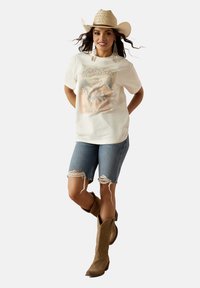 White short-sleeve t-shirt with rodeo graphic, paired with distressed denim shorts and brown cowboy boots. Cowboy hat and earrings complete the look.