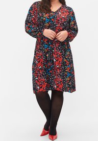 Schwarzes Blumenkleid mit roten und blauen Blumen, langen Ärmeln, V-Ausschnitt und gerafftem Taillenbereich. Kombiniert mit schwarzen Strumpfhosen und roten Absätzen.