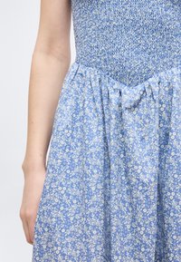 Φόρεμα μπλε με σφηκοφωλιά στο πάνω μέρος και σούρα στη μέση, με λευκό λουλουδάτο μοτίβο, φορεμένο από ένα άτομο που δείχνει τον βραχίονά του και το χέρι του.