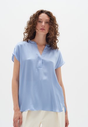 Bluză scurtă cu mâneci scurte, de culoare albastru deschis, cu decolteu în V. Textură netedă, croială lejeră și slituri laterale pentru confort. Materialul are un ușor luciu.