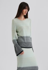 Robe en tricot vert menthe avec des accents noirs. Présente un motif texturé en diamant et des manches longues avec un ourlet inférieur évasé.