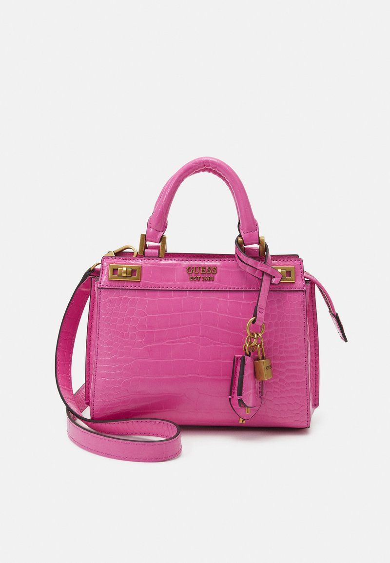 Sac à main rose vif avec texture crocodile, poignée supérieure, bandoulière amovible, ferrures dorées, charme en forme de cadenas et logo "GUESS" sur le devant.