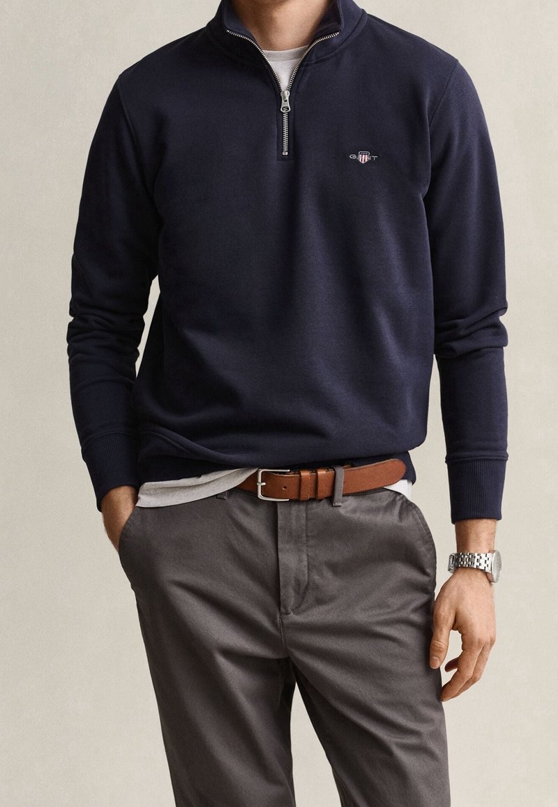 Homme portant un pull marine à fermeture éclair quart, pantalon gris avec ceinture marron et montre-bracelet argentée, debout les mains dans les poches devant un fond uni.