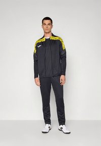 Črna športna jakna z rumenimi poudarki in logotipom, kombinirana z ujemajočimi črnimi hlačami in belimi čevlji, na enobarvnem ozadju.