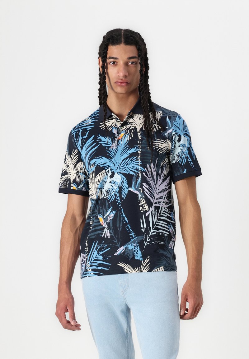 Polo shirt bleu marine avec un imprimé tropical de palmiers et d'oiseaux. Manches courtes, en coton doux, et col classique. Accents bleu clair visibles.