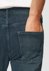 Jeans in denim blu scuro con una finitura testurizzata, dotati di una tasca laterale e dettagli di cucitura visibili in vita e ai bordi delle tasche.