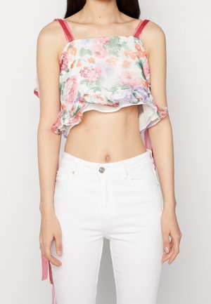 Femme portant un crop top à fleurs avec volants et bretelles roses, associé à un jean blanc taille haute, sur un fond clair uni.