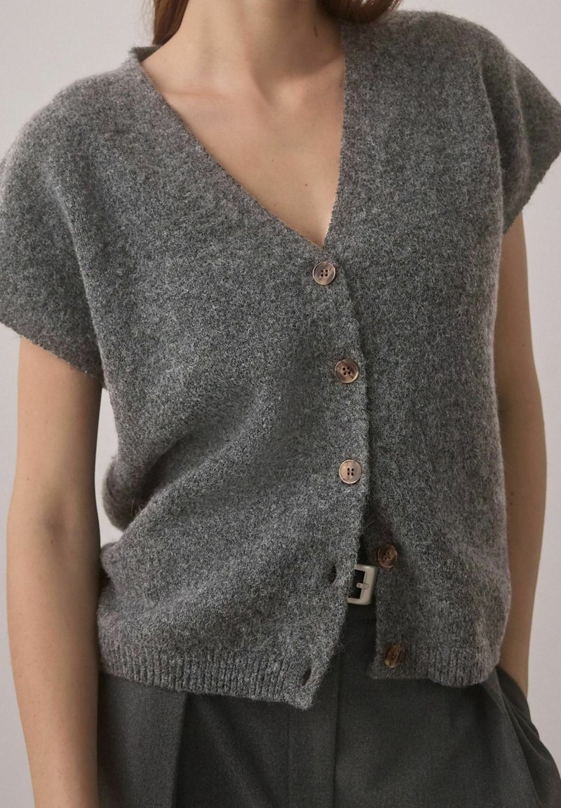 Cardigan en laine gris à manches courtes avec quatre boutons, porté sur un pantalon foncé avec une boucle de ceinture blanche visible.