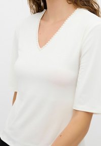 Top blanco de manga corta con escote en V adornado con una fila de pequeñas perlas. Tejido suave y liso con un diseño ajustado.
