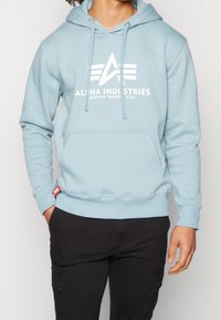 Felpa con cappuccio azzurro chiaro con una tasca frontale, caratterizzata da un logo bianco e testo. Realizzata in materiale morbido, ha un cappuccio con coulisse e polsini a coste.