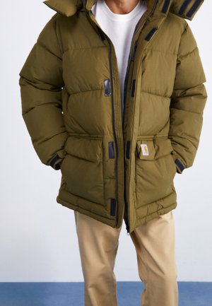 Personne portant une veste matelassée vert olive avec capuche, pantalon beige et chemise blanche, debout devant un fond uni.