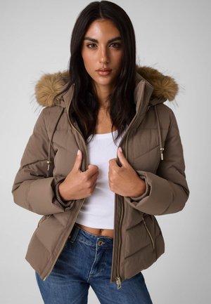 Femme portant une doudoune marron avec une capuche en fourrure par-dessus un crop top blanc et un jean bleu, tenant le devant de la veste avec les deux mains.