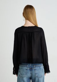 Donna con lunghi capelli lisci che indossa una blusa nera con merletto e jeans chiari a vita alta, vista di spalle contro uno sfondo semplice.