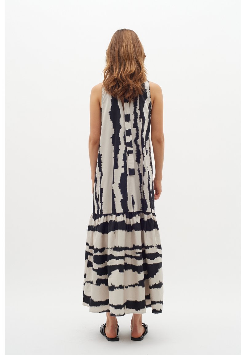 Femme aux cheveux ondulés mi-longs portant une robe maxi sans manches à motif noir et blanc et des sandales noires, de dos sur fond blanc.