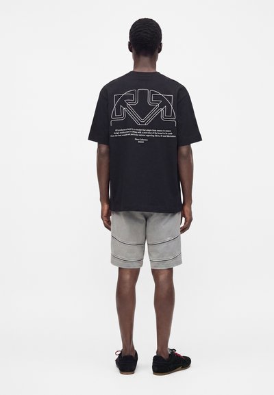Άντρας με πλάτη, φοράει μαύρο T-shirt με λευκά γραφικά βέλη και κείμενο στην πλάτη, γκρι σορτς έως το γόνατο και μαύρα παπούτσια.