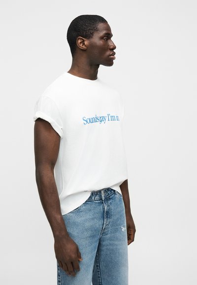 Άνδρας με λευκό T-shirt με γυρισμένα μανίκια και μπλε κείμενο, φορώντας ανοιχτό μπλε τζιν, στέκεται πλευρικά σε λευκό απλό φόντο.