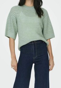 Ljusgrön stickad tröja med en lös, boxig passform, som har ett texturerat, öppet vävmönster och ribbade ärmslut, kombinerad med mörkblå vida jeans.