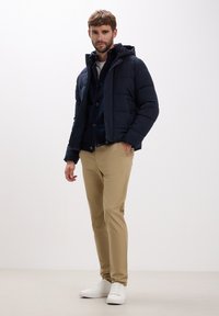 Giubbotto imbottito blu navy con cappuccio, camicia scura con bottoni sotto, pantaloni beige e sneakers bianche. Texture liscia e design casual.