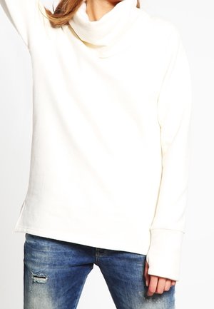 Osoba ubrana w biały top z kapturem i długim rękawem oraz niebieskie podarte dżinsy, unosząca jedną rękę, a druga jest opuszczona przy boku.