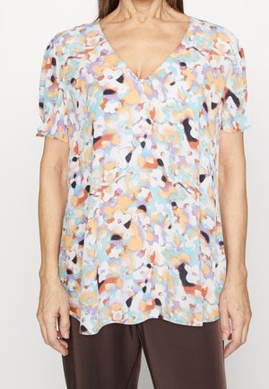 Femme portant un chemisier à manches courtes et col en V avec un motif floral abstrait multicolore et un pantalon marron foncé.