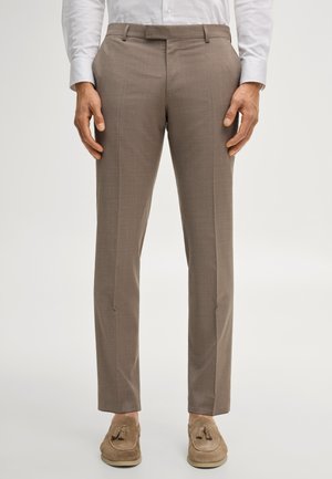 Uomo che indossa pantaloni eleganti marroni slim-fit con piega centrale, abbinati a mocassini beige con nappine, in piedi davanti a uno sfondo neutro.