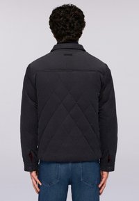 Svart vadderad jacka med diamantmönster, med krage och tryckknappar vid ärmarna, matchad med blå jeans.