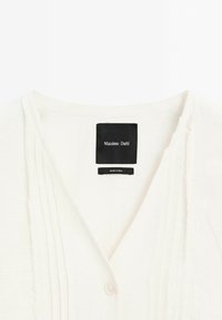 Biała lniana bluzka z dekoltem w kształcie litery V, z pionowymi fałdami i pojedynczym zapięciem na guzik. Metka głosi "Massimo Dutti", wyprodukowana w Indiach.