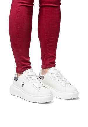 White leather sneakers with a thick white sole, featuring black accents on the heel and a logo on the side. Worn with red skinny jeans.