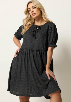 Blondynka w luźnej czarnej sukience o fakturowanej tkaninie, z bufiastymi rękawami i detalami wiązanymi przy dekolcie, pozująca na jednolitym tle.