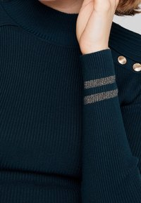 Suéter de punto acanalado en navy con cuello alto y mangas largas, que presenta tres detalles decorativos de rayas doradas en una manga y botones dorados en el hombro.