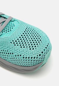 Scarpa atletica in mesh di un azzurro chiaro, con una superficie testurizzata e un motivo traspirante, dettagli grigi sul tallone e sulla linguetta, e forma arrotondata.