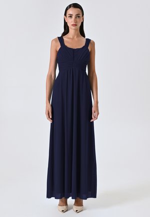 Robe maxi bleu marine avec un buste plissé, des bretelles larges et une jupe fluide. Tissu lisse avec un éclat subtil. Associée à des chaussures de couleur claire.