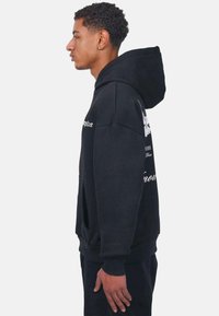 Sort hættetrøje med kængurulomme, oversized pasform, ribbede cuffs og kant, med grafiktrykte motiver på forsiden og bagsiden.