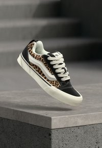 Ασπρόμαυρο αθλητικό παπούτσι με πλευρές σε λεοπάρ σχέδιο και λευκές κορδόνια, να αιωρείται πάνω από μια τσιμεντένια άκρη με θολές σκάλες στο φόντο.