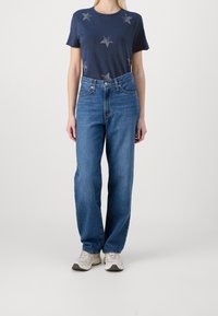 Camiseta navy con patrones de estrellas plateadas, combinada con jeans de tiro alto en azul claro. El modelo lleva zapatillas claras para un look informal.