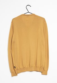 Pull moutarde à coupe décontractée, manches longues et poignets côtelés. Texture lisse, suspendu à un crochet noir sur un fond blanc.