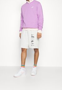 Fioletowa bluza z wysokim kołnierzem, biały logo Nike. Jasnoszare spodenki z wieloma logo Nike. Białe buty z różowymi akcentami i skarpetki w paski w kolorach tęczy.