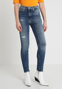 Vaqueros ajustados de talle alto en un denim azul medio, con detalles desgastados y ligeras marcas de uso. Combinados con botas blancas de tacón puntiagudo.