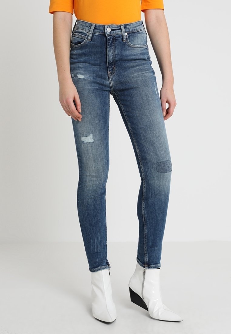Vaqueros ajustados de talle alto en un denim azul medio, con detalles desgastados y ligeras marcas de uso. Combinados con botas blancas de tacón puntiagudo.