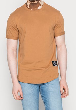 Homme portant un t-shirt marron uni à manches courtes avec un patch logo CK noir et un jean bleu clair, debout devant un fond uni.