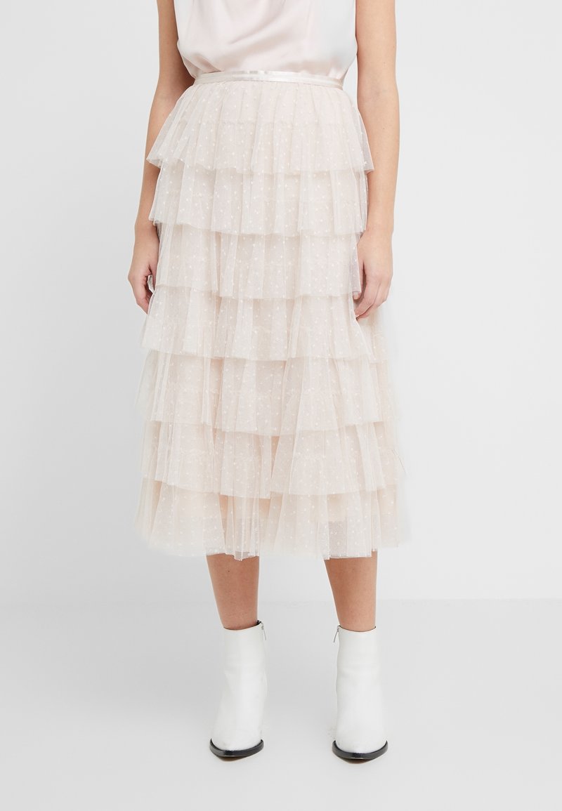 Femme portant une jupe midi en tulle crème à motifs à pois et des bottines blanches, debout devant un fond clair uni.