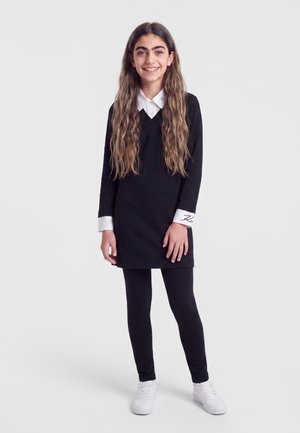 Fille souriante aux longs cheveux ondulés, portant une robe noire avec col et poignets blancs, des leggings noirs et des baskets blanches, debout sur fond blanc.