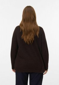 Persoon met lang bruin haar, gekleed in een donkerbruine trui en donkere broek, staande met de rug naar de camera tegen een effen witte achtergrond.