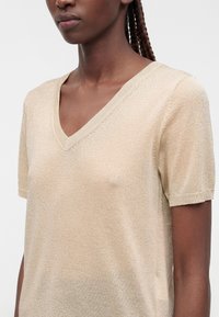 Femme portant un haut en maille beige clair à manches courtes avec col en V, visible des épaules au milieu du torse, avec les cheveux tressés tirés en arrière.