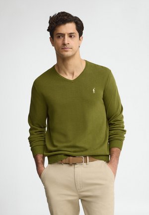 Hombre con cabello rizado y oscuro, vistiendo un suéter de cuello en V verde oliva y pantalones beige, de pie con las manos en los bolsillos, mirando de lado.