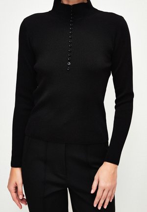 Woman wearing a fitted black long-sleeve top with a high collar and a vertical row of small black buttons down the front.