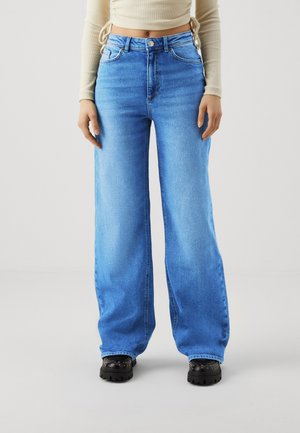 Femeie care poartă blugi albastre deschis cu picioare largi, pantofi negri cu ținte și un top bej scurt, cu mâneci lungi, stând în fața unui fundal simplu.