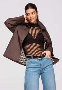 Femme posant avec une veste marron ouverte, portant un haut en dentelle noire, un jean bleu clair et une ceinture noire avec une boucle dorée, sur fond uni.