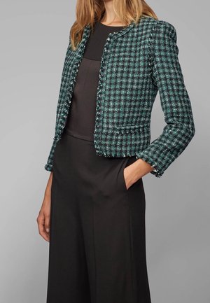 Kobieta w zielono-czarnej kratowanej krótkiej marynarce na czarnej sukience, z jedną ręką w kieszeni na szarym tle.